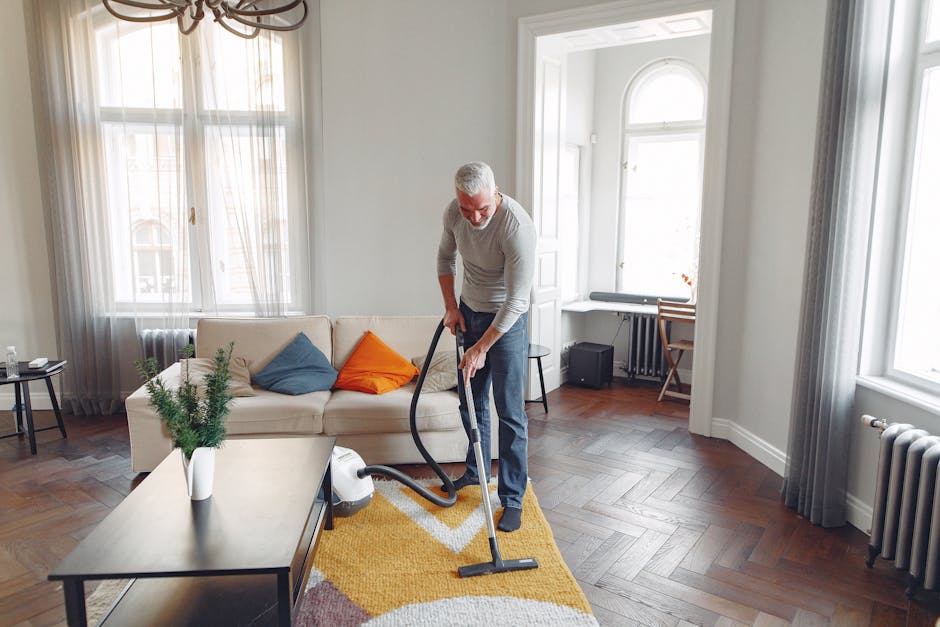 A professional cleaning specialist dressed in full protective gear, including a white coverall, gloves, face mask, and shoe covers, is performing deep carpet cleaning in a bright, modern living room.