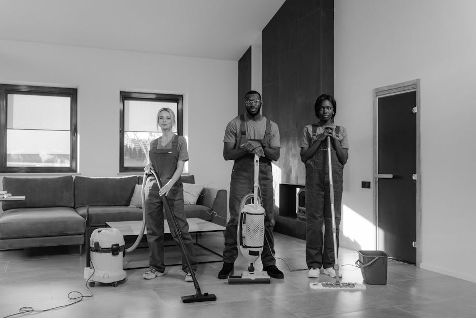 A person dressed in a grey sweater and black pants is using a mop to clean a dark grey tiled floor in a living room. The room features a large window with natural light, a grey sofa with light-colored