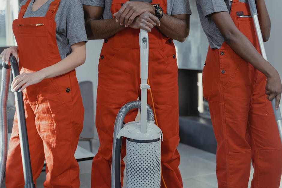 A young man with dark curly hair, dressed in an orange disposable coverall, stands indoors against a plain light grey wall. He holds two large blue plastic garbage bags, one in each hand, which are fi