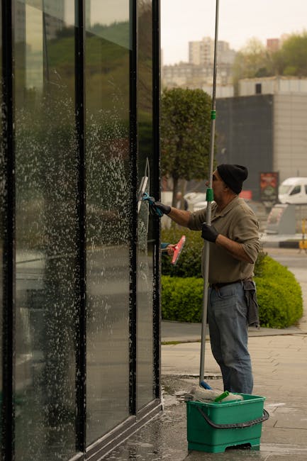 A professional window cleaning service in progress on a large modern office building with a glass façade. A suspended scaffold platform with a cleaning bucket and tools is visible, allowing access to