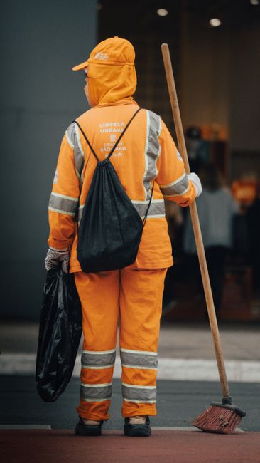 Close-up view of three professional cleaners in grey shirts and bright orange overalls, each holding cleaning tools. The person in the center is holding a vacuum cleaner with a white and black perfora